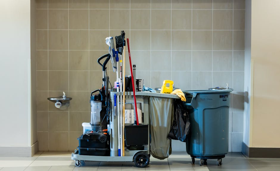 A cleaning cart positioned against a beige tiled wall in a commercial or office space, containing cleaning tools such as mop handles, spray bottles, and a vacuum cleaner. To the right, a yellow saniti